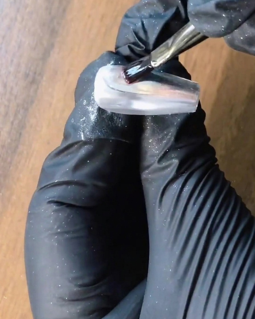 Un pincel aplicando gel de refuerzo transparente sobre una uña cromada plateada.