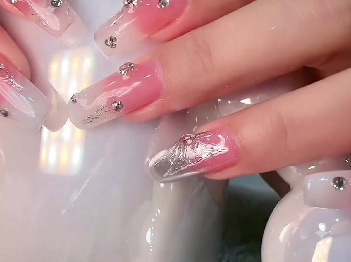 Primer plano de uñas largas cuadradas con degradado blanco lechoso y pedrería sostenidas contra una taza de cerámica blanca.