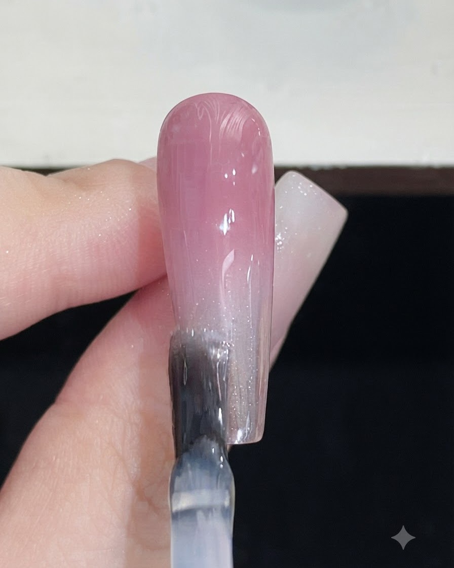 Un pincel de uñas aplicando un top coat transparente sobre una uña terminada con degradado rosa.