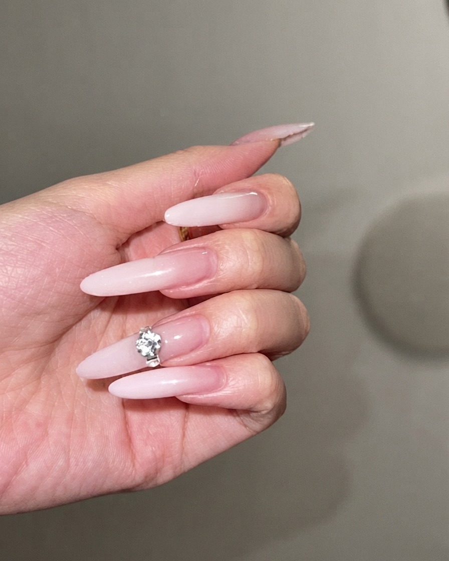 Un gran cristal brillante siendo presionado en el centro de una uña stiletto larga con degradado blanco lechoso.