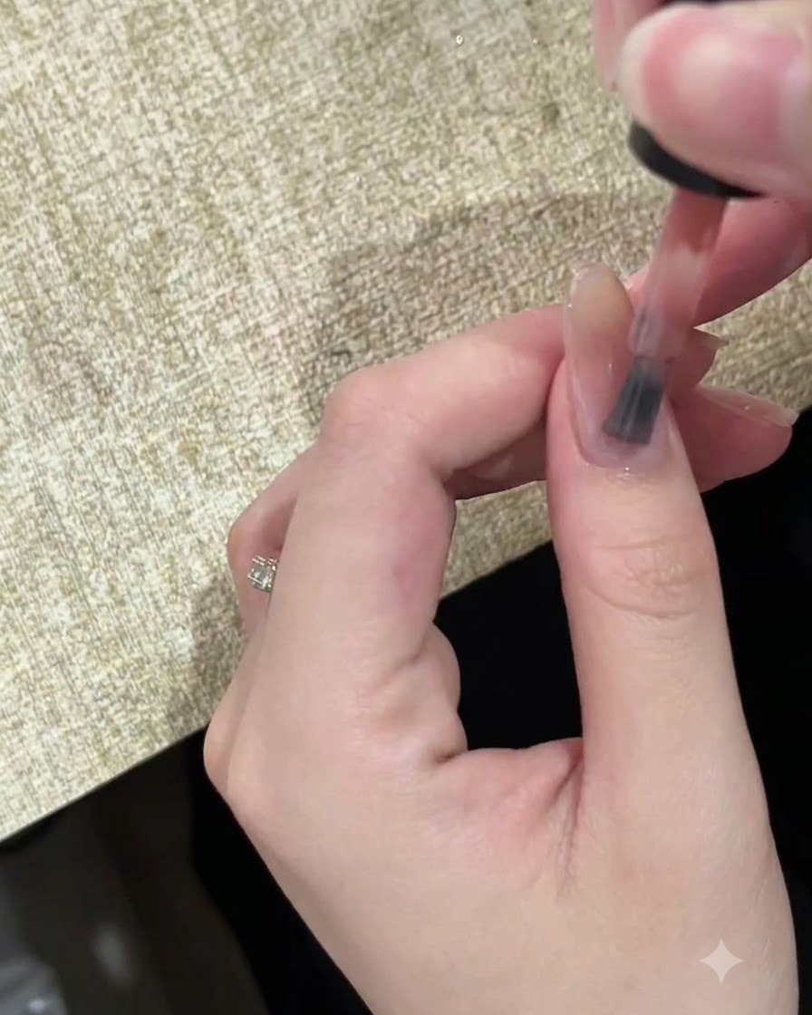 Un primer plano de un pincel aplicando un esmalte en gel translúcido color nude sobre una uña natural.