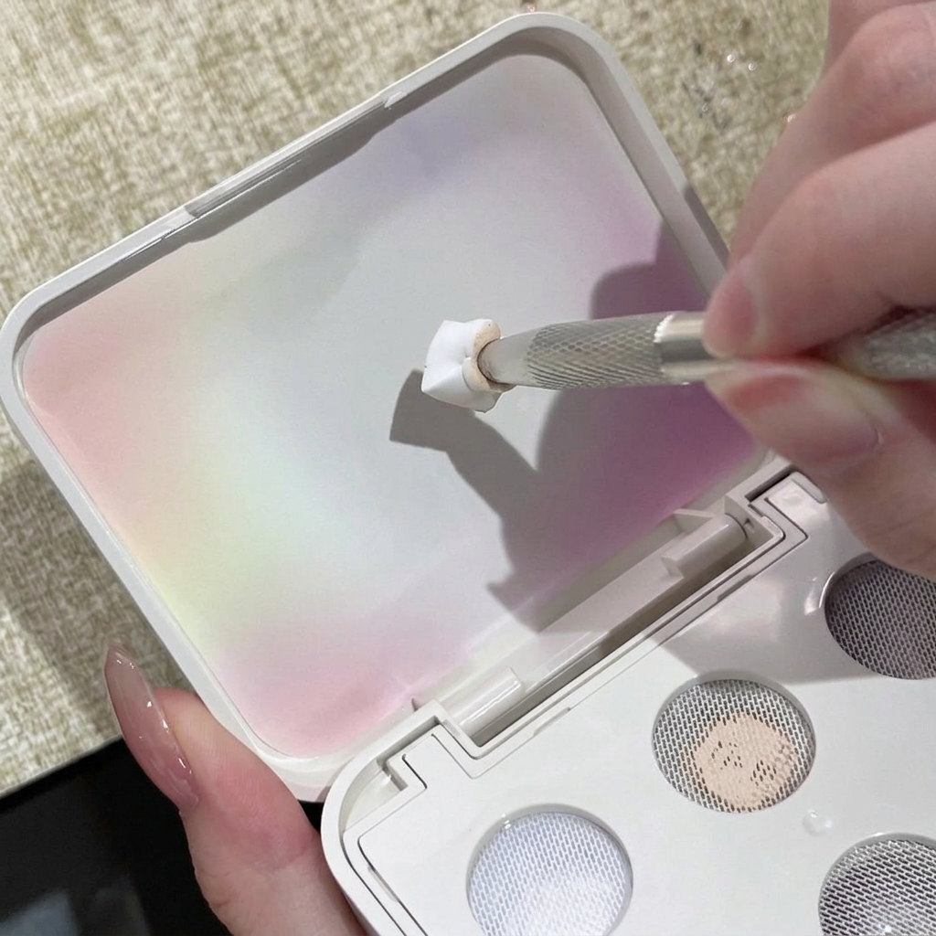 Una herramienta metálica para uñas presionando gel sólido blanco sobre la cuadrícula texturizada de la tapa de una paleta.