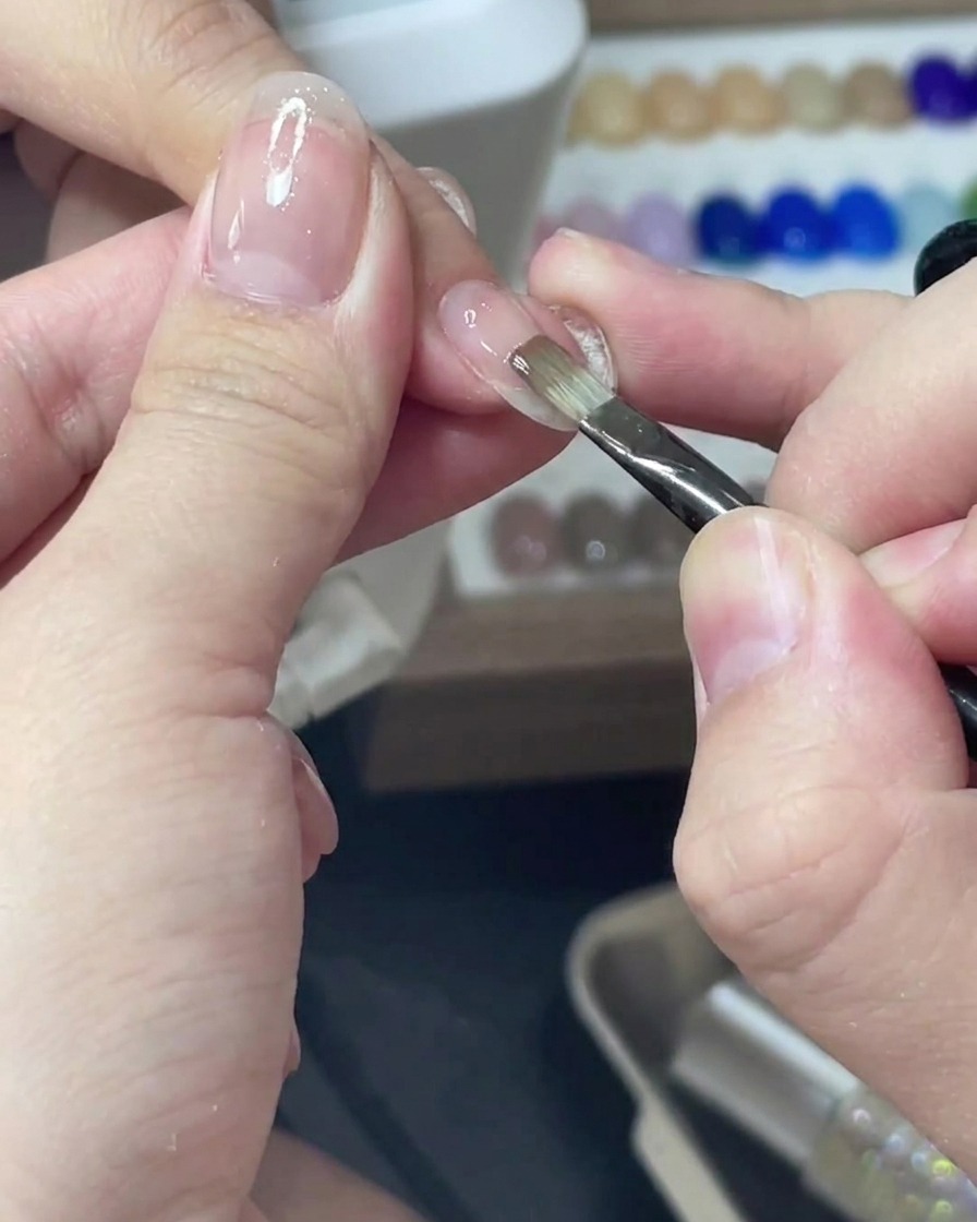 Un pincel delineador fino aplicando esmalte de gel rosa translúcido de manera uniforme sobre una uña.