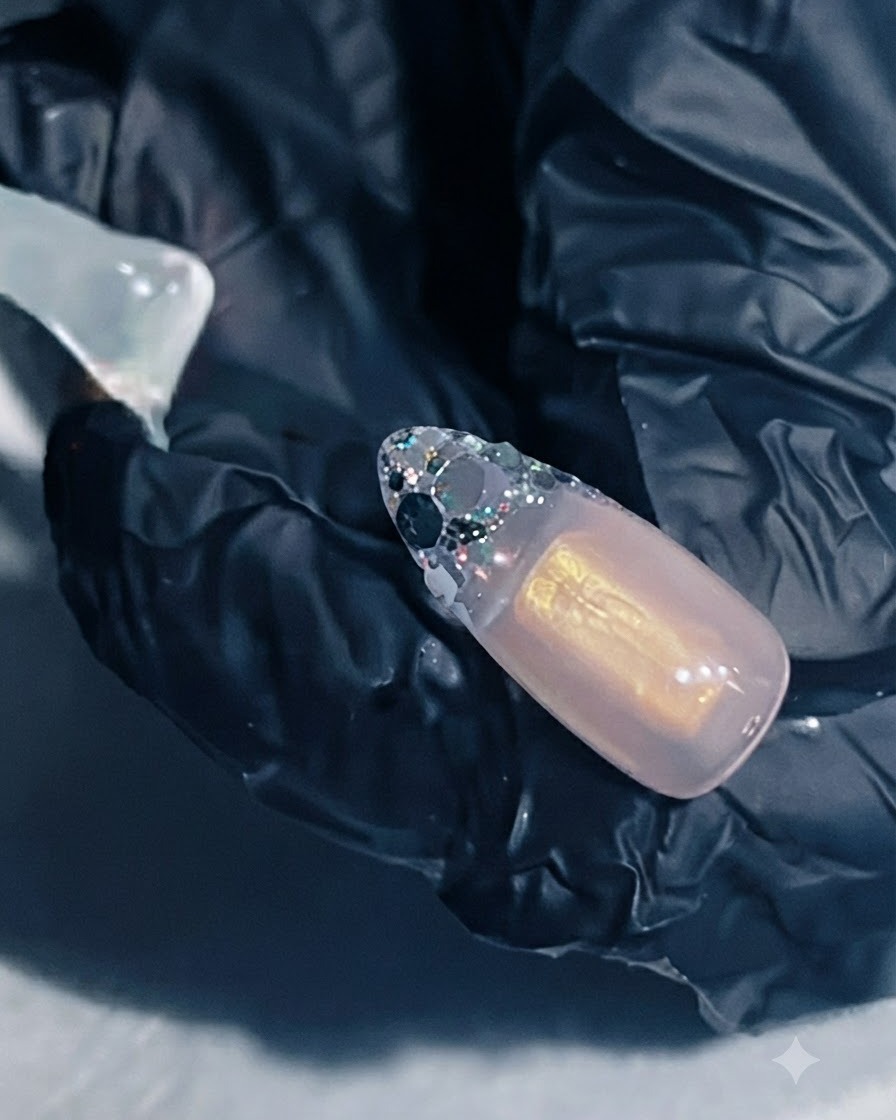 Una mano sosteniendo una extensión de uña con gel de refuerzo aplicado sobre el área francesa con glitter.