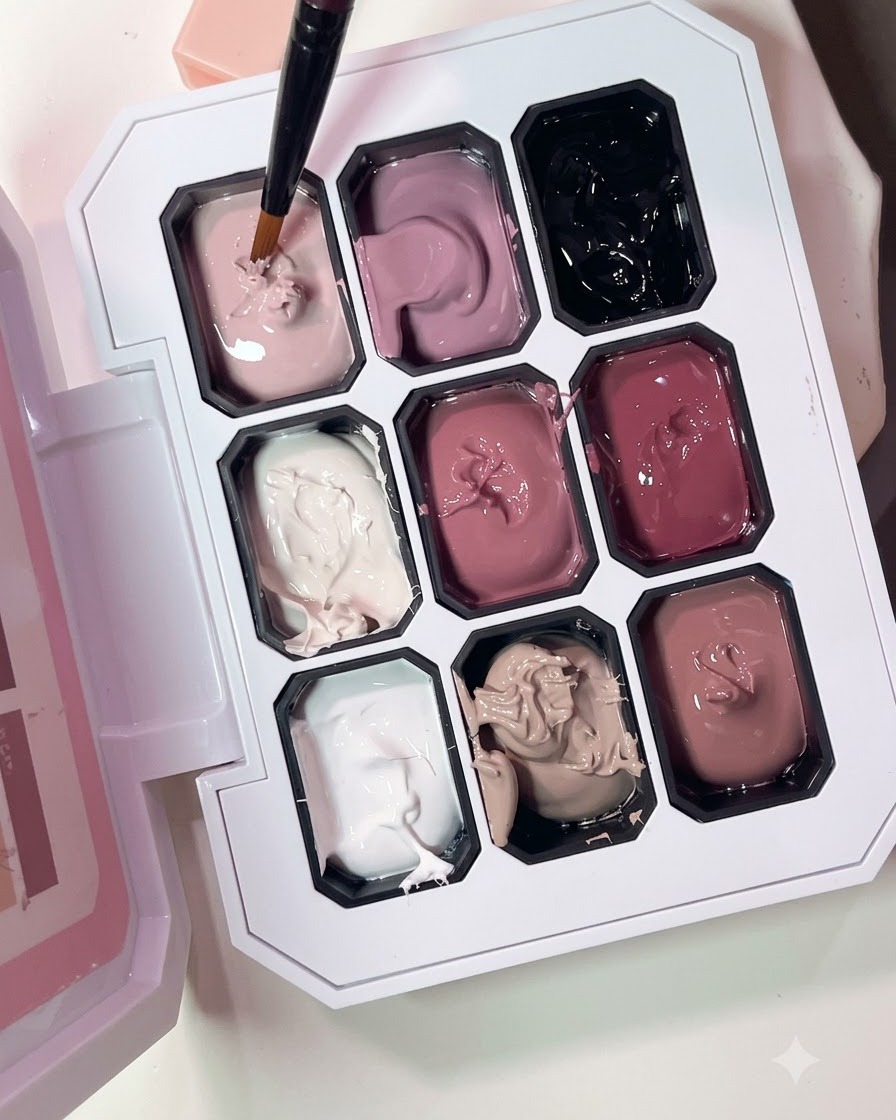 Un pincel fino tomando esmalte de gel rosa nude de una paleta de múltiples compartimentos.