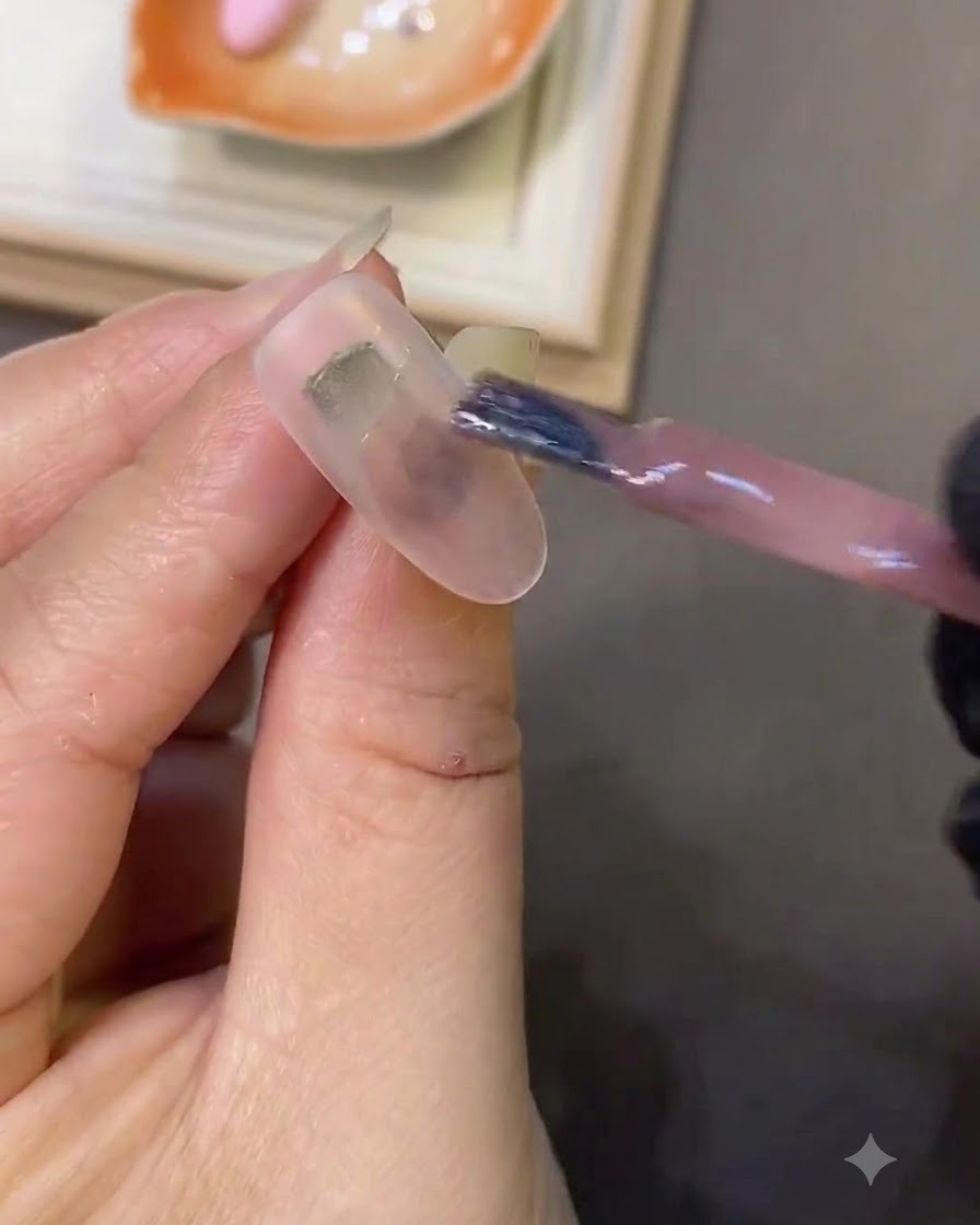 Un pincel aplicando gel ojo de gato rosa claro sobre una uña de color blanco lechoso.