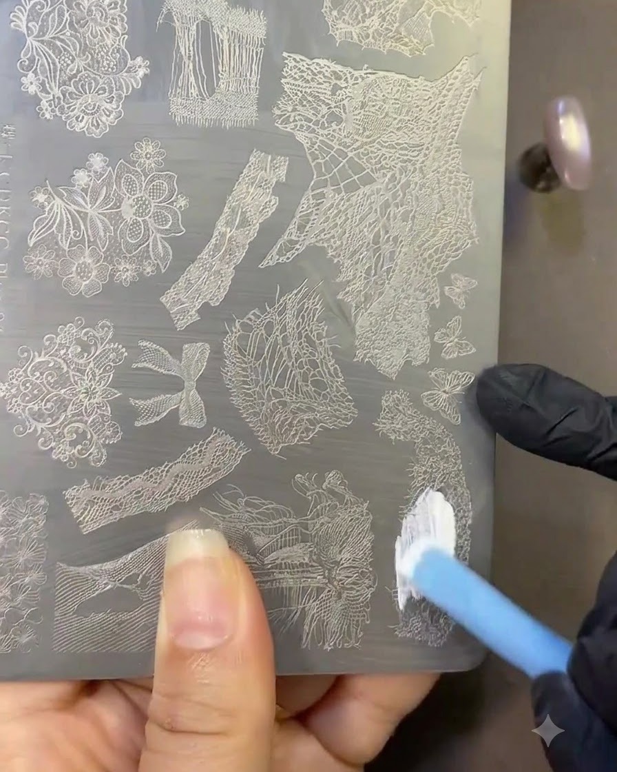 Un pincel aplicando esmalte de estampado blanco sobre una placa de metal grabada con diseños de encaje.