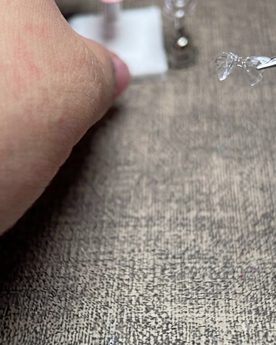 Un par de pinzas sosteniendo un delicado lazo 3D transparente justo encima de un soporte de exhibición de uñas postizas preparado.