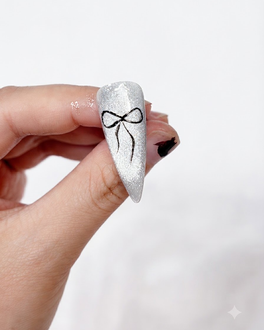 Un diseño completo de arte de uñas con lazo negro pintado limpiamente y fijado sobre un fondo cat-eye plateado brillante, sostenido entre los dedos.