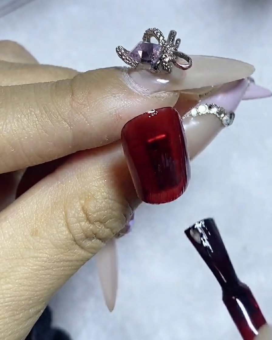 Un pincel delineador aplicando una línea precisa de pegamento para gemas sobre la superficie de una uña con esmalte rojo oscuro.