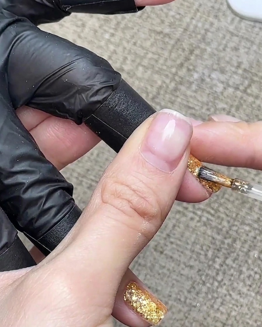 Una pincelada detallada aplicando gel marrón ámbar translúcido en la punta de una uña con purpurina dorada.