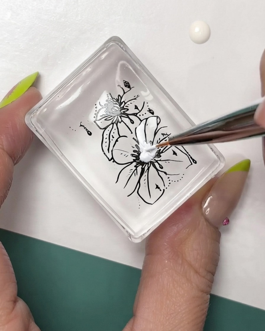 Un pincel delineador de arte de uñas fino agregando detalles blancos al estampado floral con contorno negro en una almohadilla de sello transparente.