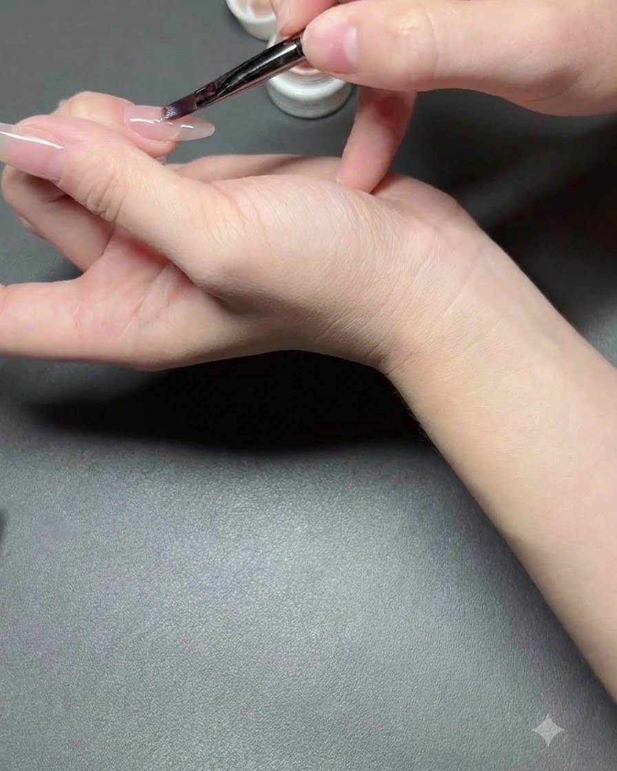 Un pincel de uñas aplicando una capa delgada de esmalte semitransparente nude en una uña.