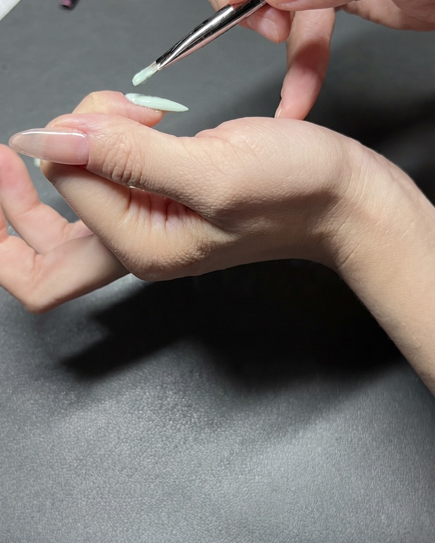 Una vista detallada de un pincel de uñas aplicando esmalte de gel verde menta con precisión en un dedo índice.