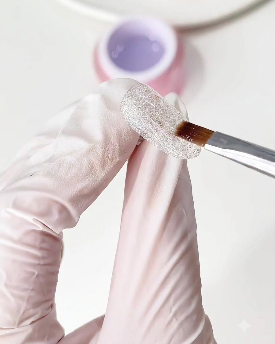 Un pincel aplicando una capa transparente de gel base sobre la base de uña brillante de diamante triturado.