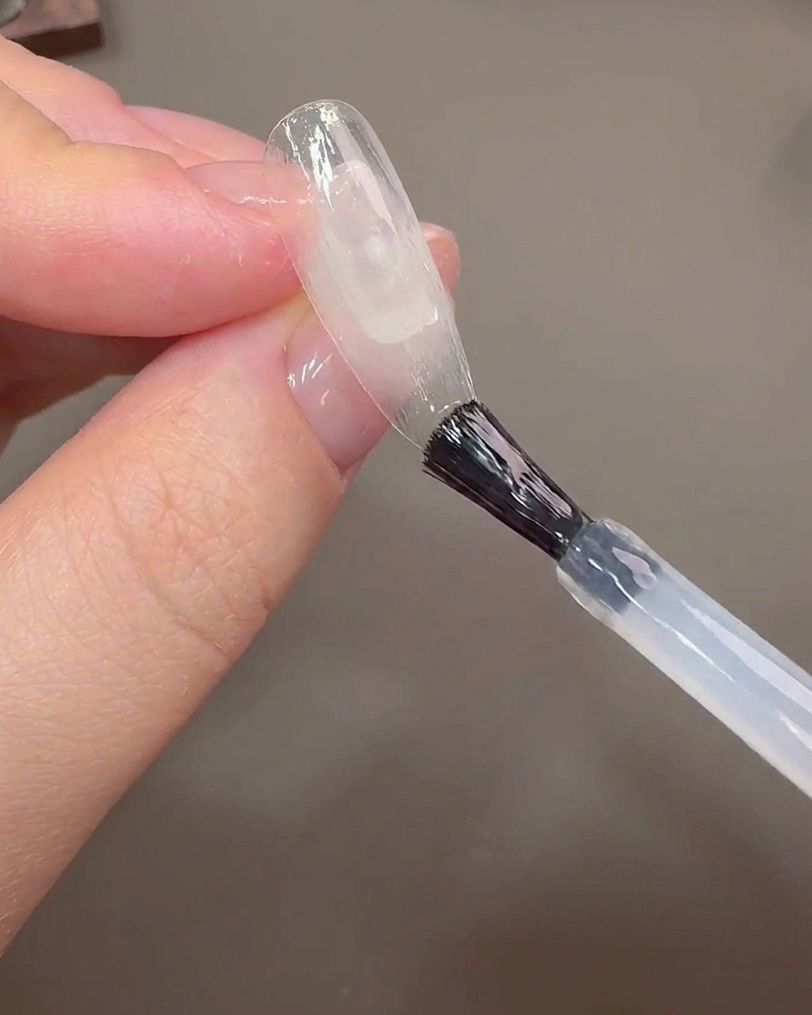 Un pincel de uñas aplicando gel de construcción sobre un tip de uña transparente para cubrir la línea de crecimiento.