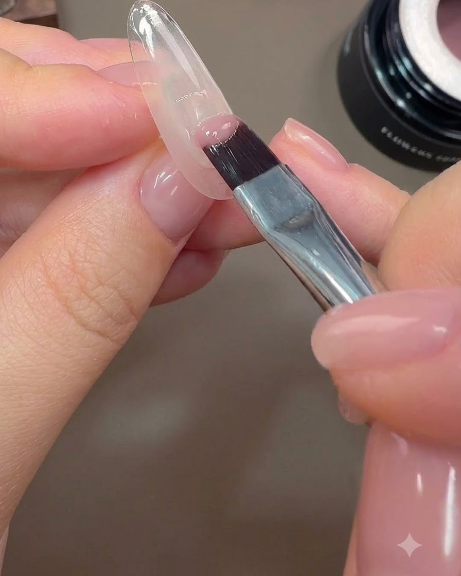 Un pincel de uñas aplicando una cobertura parcial de gel de color rosa nude en el borde posterior de una uña.