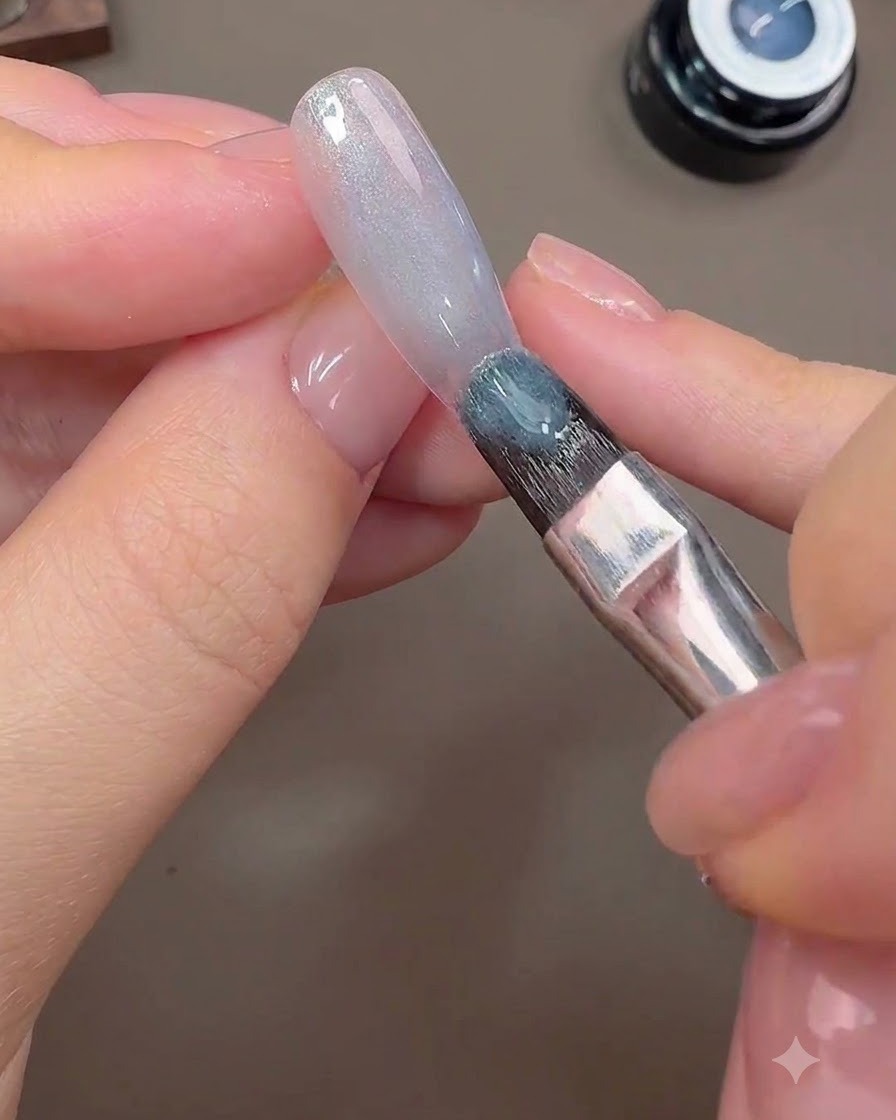 Un pincel aplicando gel ojo de gato gris-azulado sobre la superficie de la uña para formar el fondo galáctico.