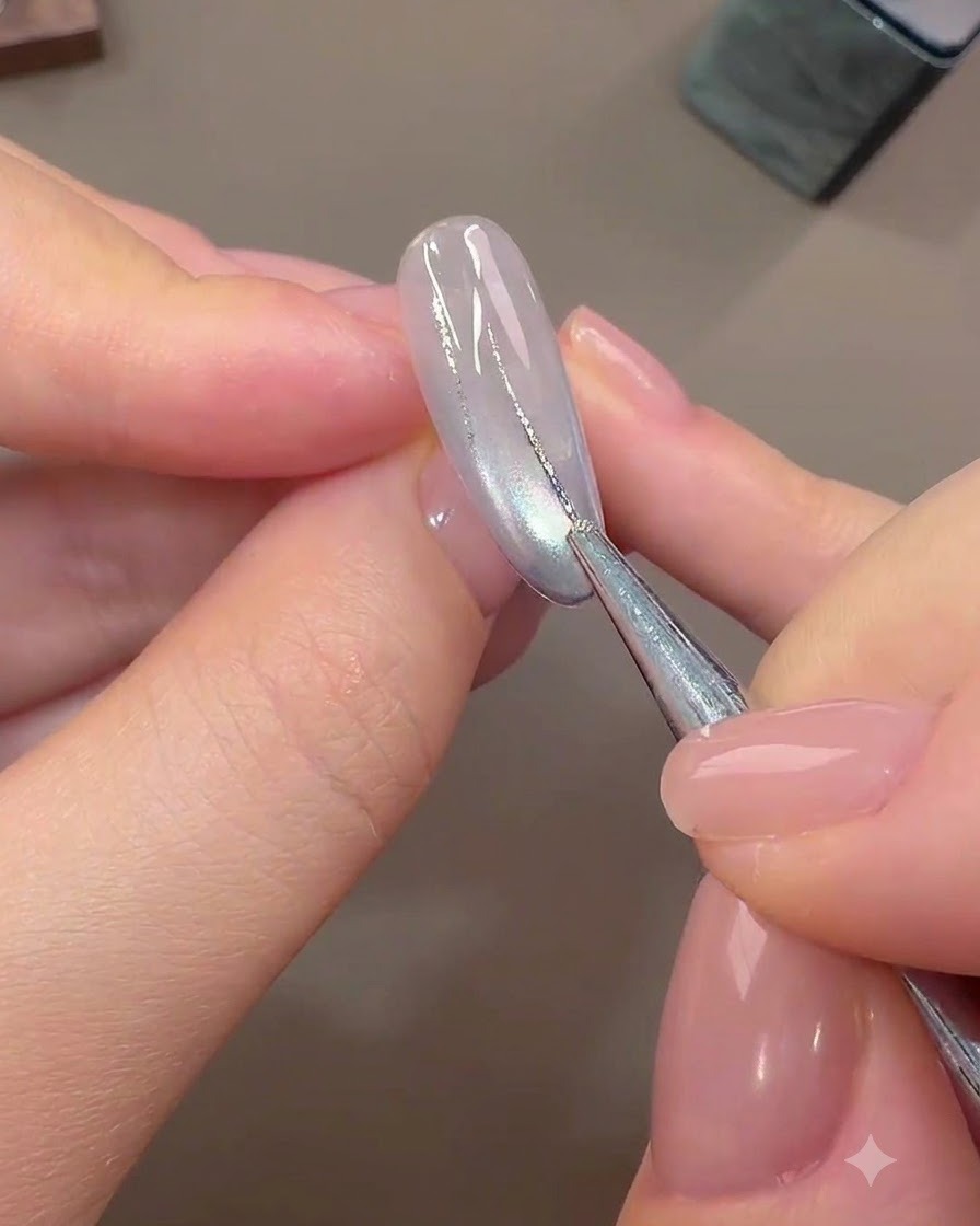 Un pincel fino de nail art dibujando líneas abstractas finas con gel de lámina plateada sobre una uña base curada de color claro.