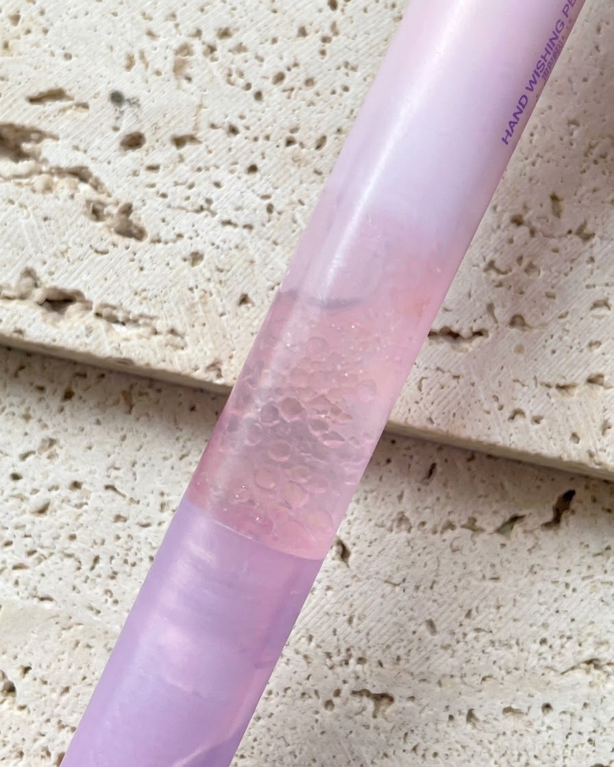 Un lápiz de aceite para cutículas de esencia perlada con degradado rosa sostenido contra un fondo texturizado.