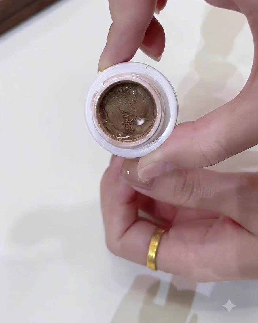 Un primer plano de un frasco pequeño abierto lleno de gel cat eye color oro champaña oscuro sostenido por una mano.