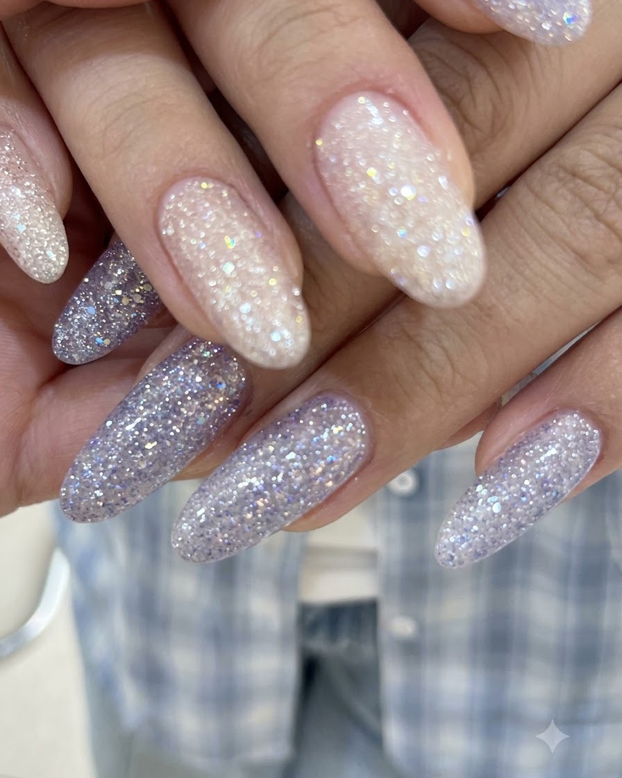Un primer plano de manos con los dedos entrelazados que muestran uñas de gel con glitter brillante en tonos nude rosado y morado.