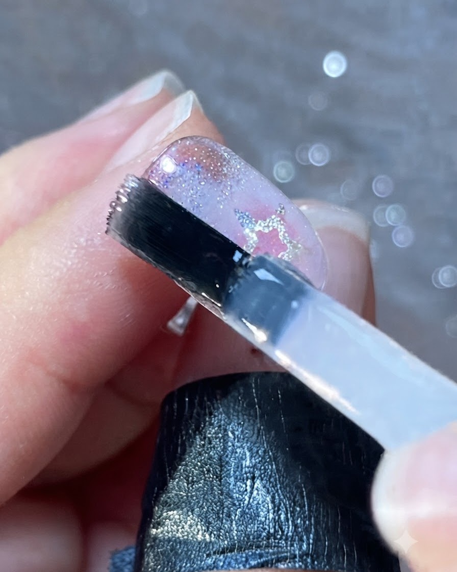 Un pincel de uñas aplicando una capa uniforme de esmalte de brillo transparente sobre unas uñas decoradas.
