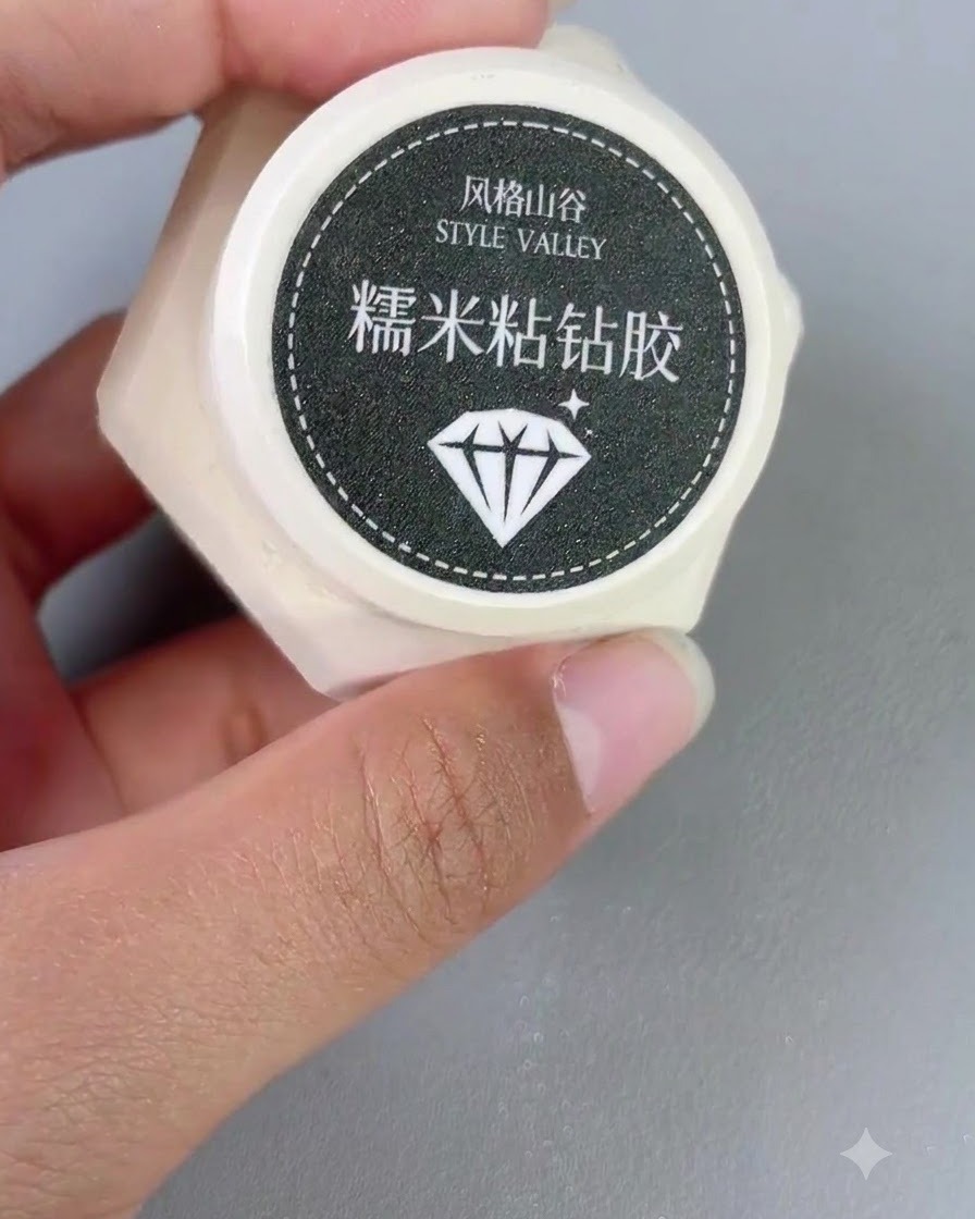 Un pequeño recipiente de gel adhesivo de diamantes sostenido en una mano junto a un pincel de uñas.
