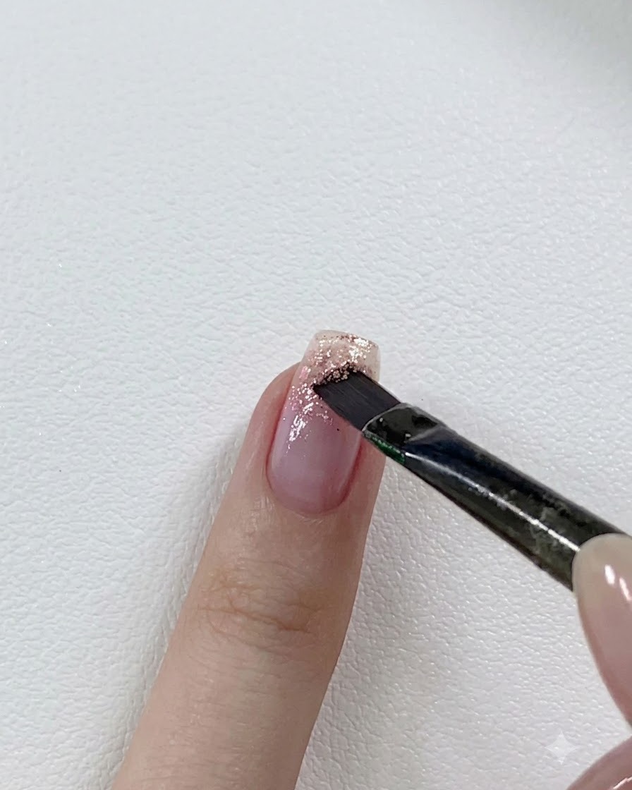 Un pincel de uñas aplicando una capa de gel de purpurina rosa más oscuro sobre la punta de una uña parcialmente con purpurina.