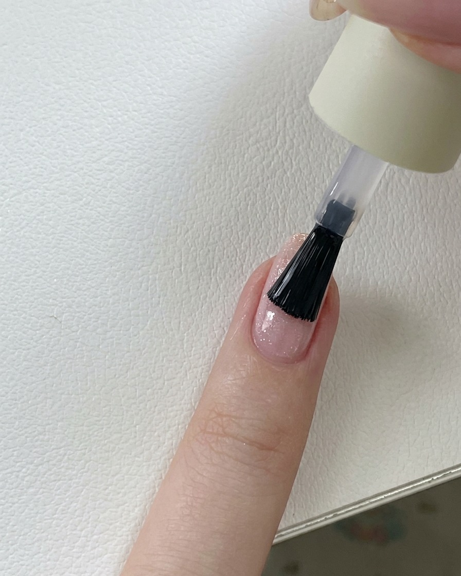 Un pincel aplicando una capa superior transparente y brillante sobre una uña de gel con purpurina rosa.