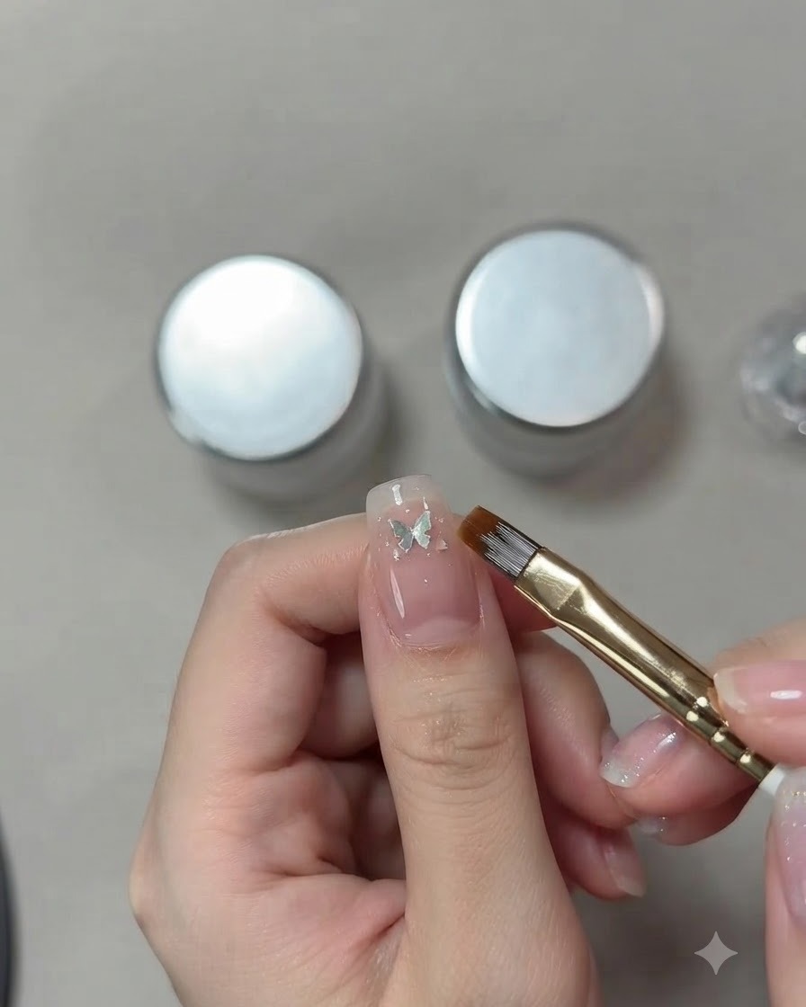 Primer plano detallado de la mano de una artista de uñas utilizando un pincel fino para colocar una pequeña lentejuela de mariposa plateada sobre una uña ojo de gato rosa.