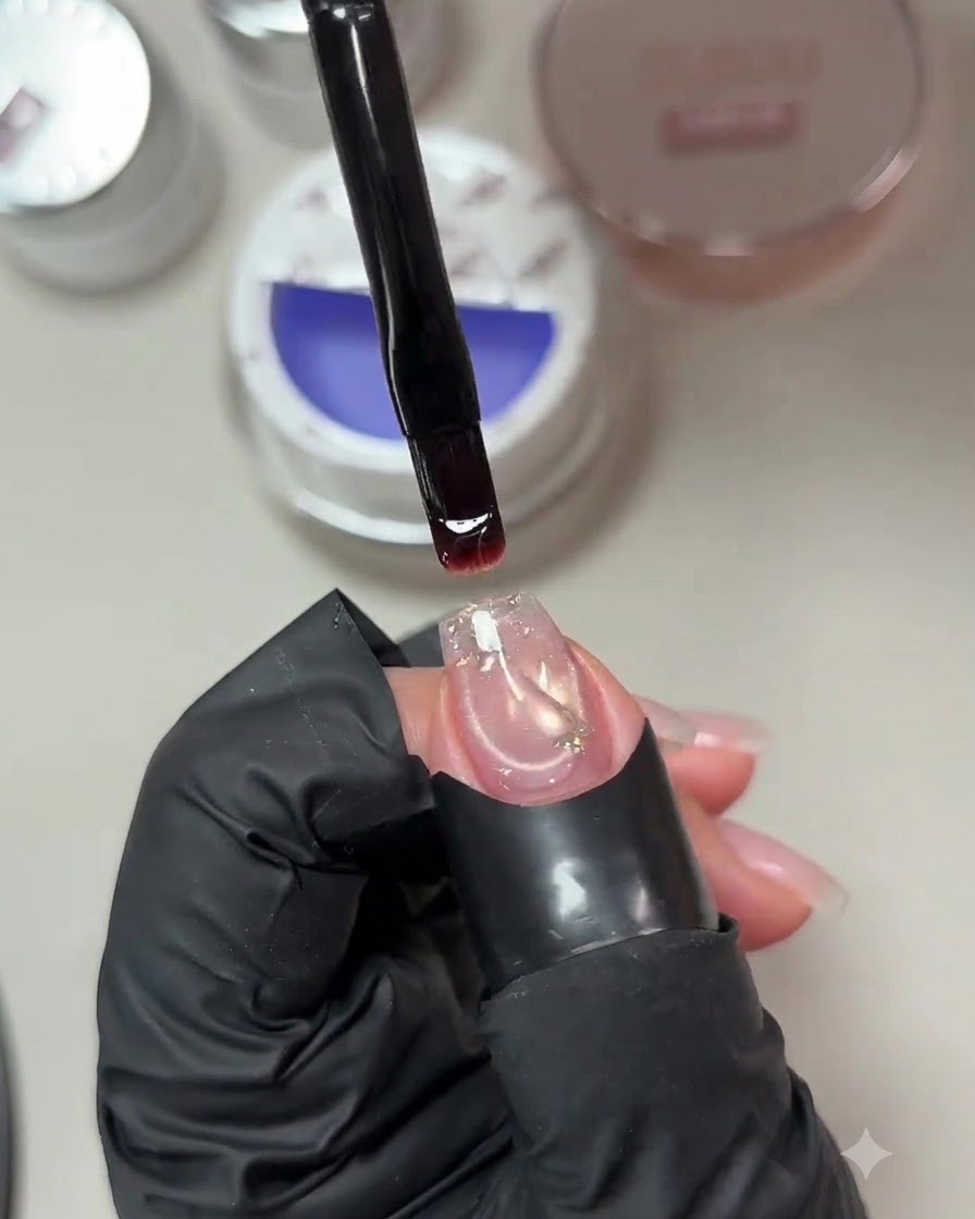 Pincel sosteniendo una gota de gel constructor azul violáceo pálido sobre una uña preparada con una lentejuela encapsulada.