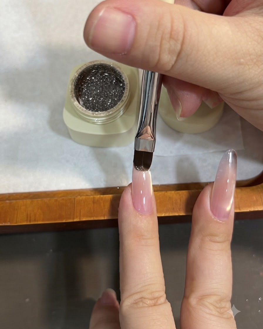 Un pincel plano aplicando gel de purpurina marrón brillante en la punta de una uña almendrada, deslizándolo hacia abajo para crear un efecto ombre.