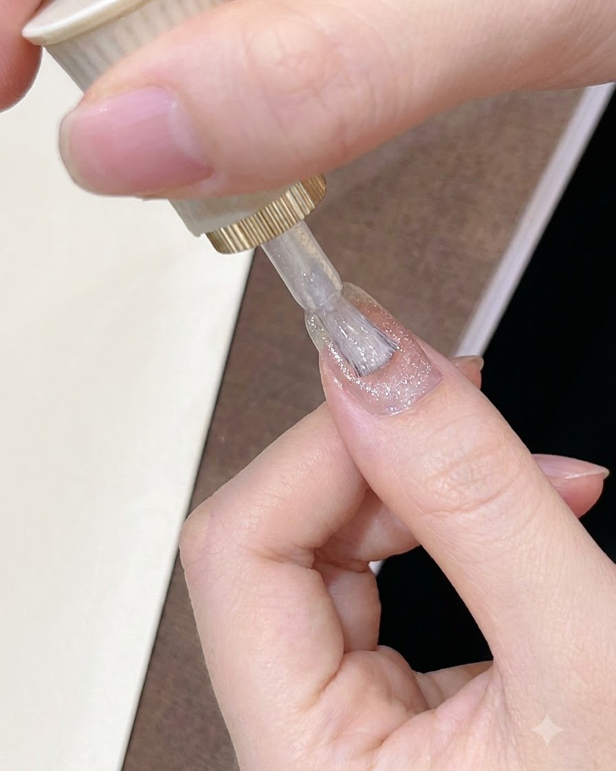 Un pincel aplicando un esmalte en gel brillante y translúcido de tono marrón rosado sobre la superficie de la uña.