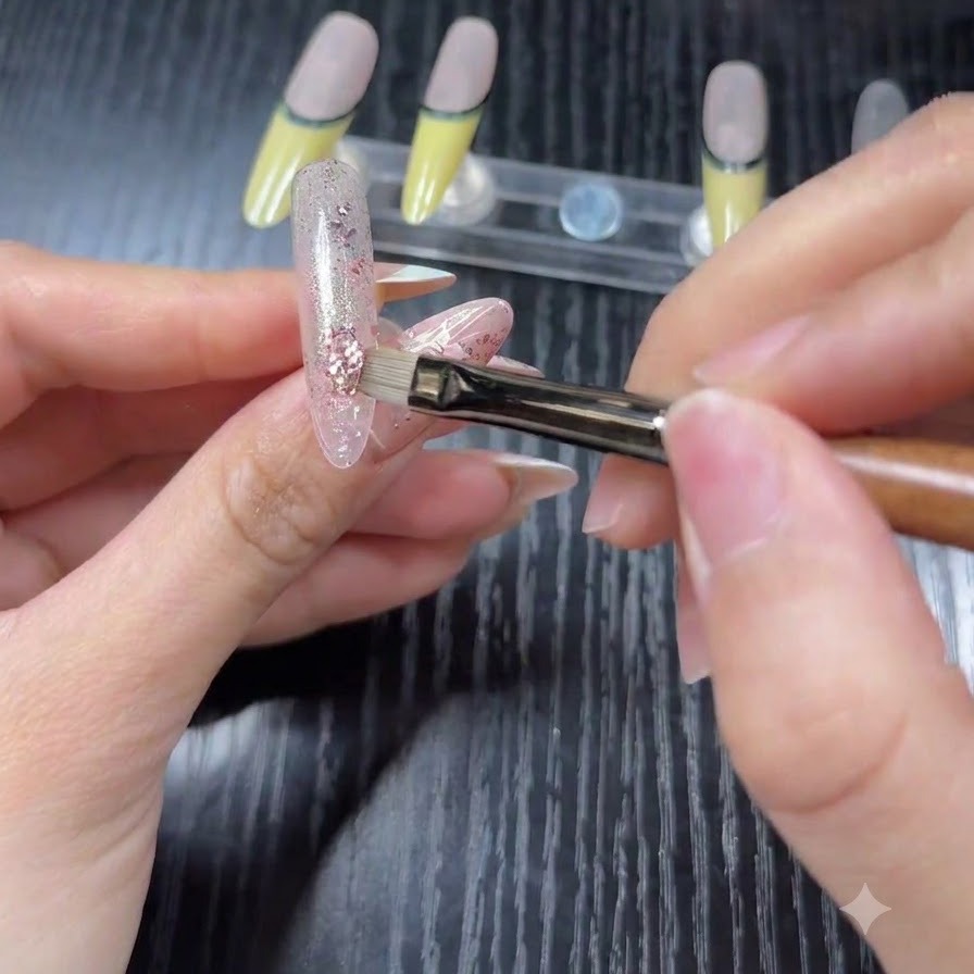 Un primer plano de un pincel aplicando escamas de purpurina holográfica rosa y plateada sobre una base translúcida en una punta de uña de práctica.