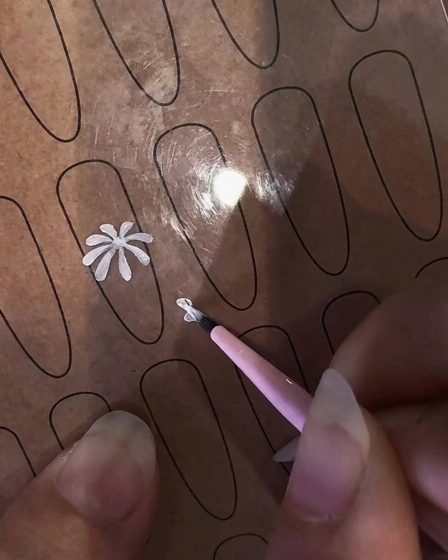 Un primer plano de una mano sosteniendo un pincel delineador fino para pintar un pétalo de margarita blanca en una plantilla de práctica transparente.