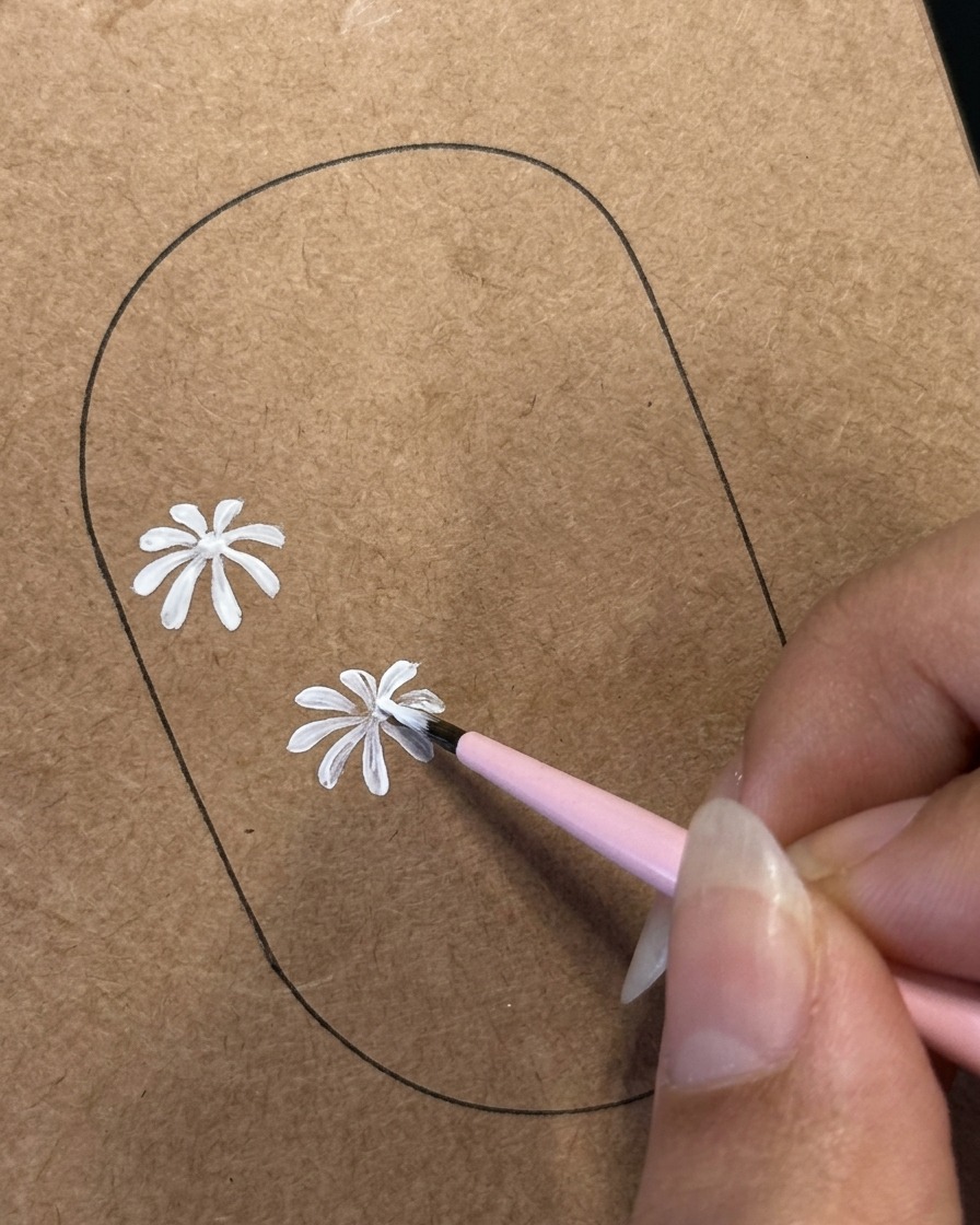 Un pincel delineador fino detallando pétalos blancos en un segundo diseño de margarita pintado sobre una plantilla de práctica marrón.