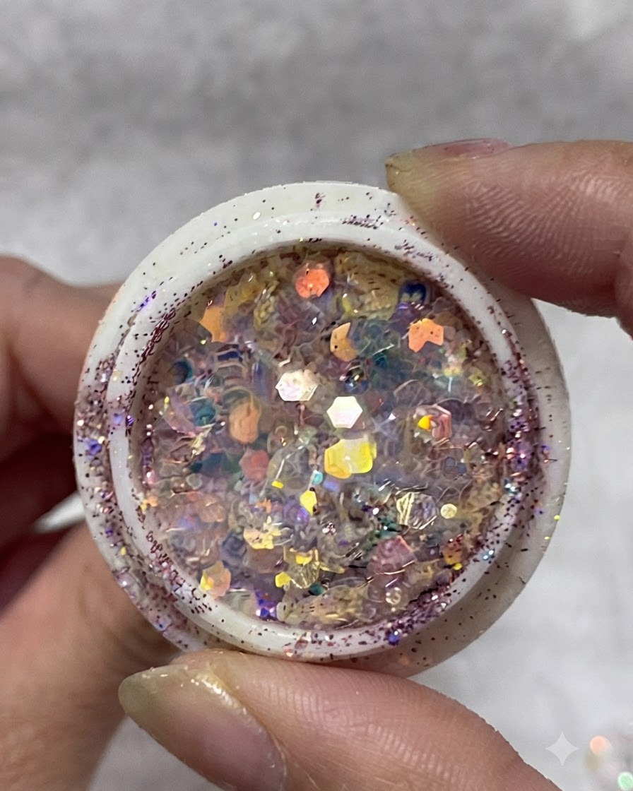 Un frasco lleno de escamas de purpurina iridiscente multicolor, lentejuelas y pequeñas estrellas sostenido entre los dedos.