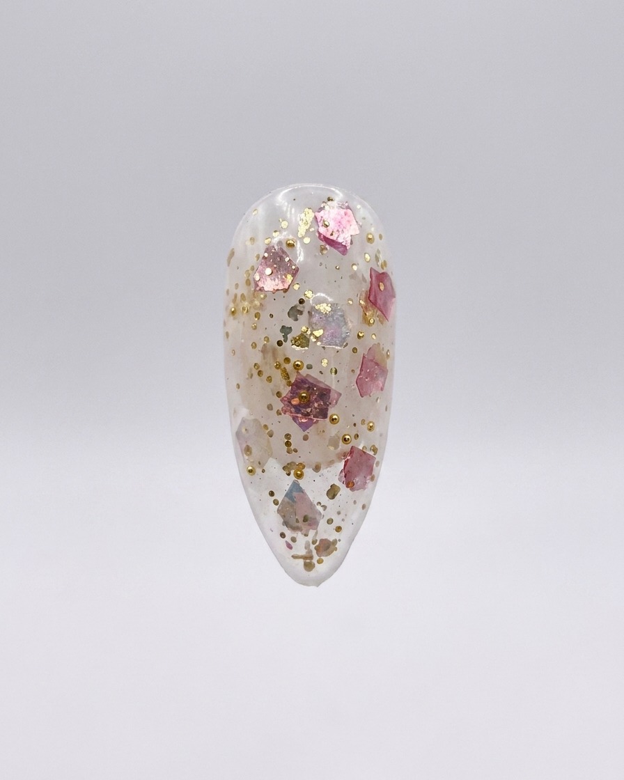 Una uña postiza transparente con escamas de purpurina rosa y dorada incrustadas, mostrada aislada sobre un fondo gris claro.