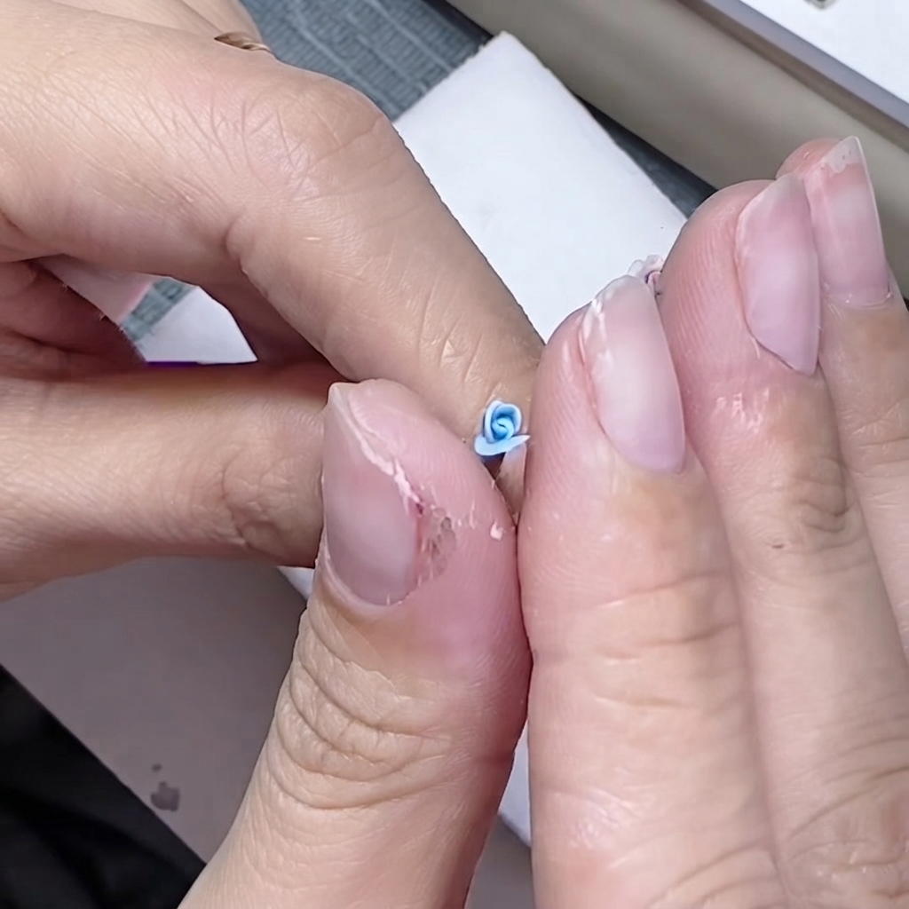 Dedos pellizcando delicadamente la base de un pequeño pétalo azul alrededor de un núcleo de rosa esculpido.