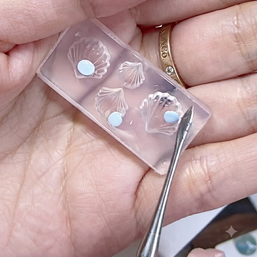 Un molde de silicona sostenido en una mano con perlas de gel azul aplanadas y una herramienta de esculpido cerca.