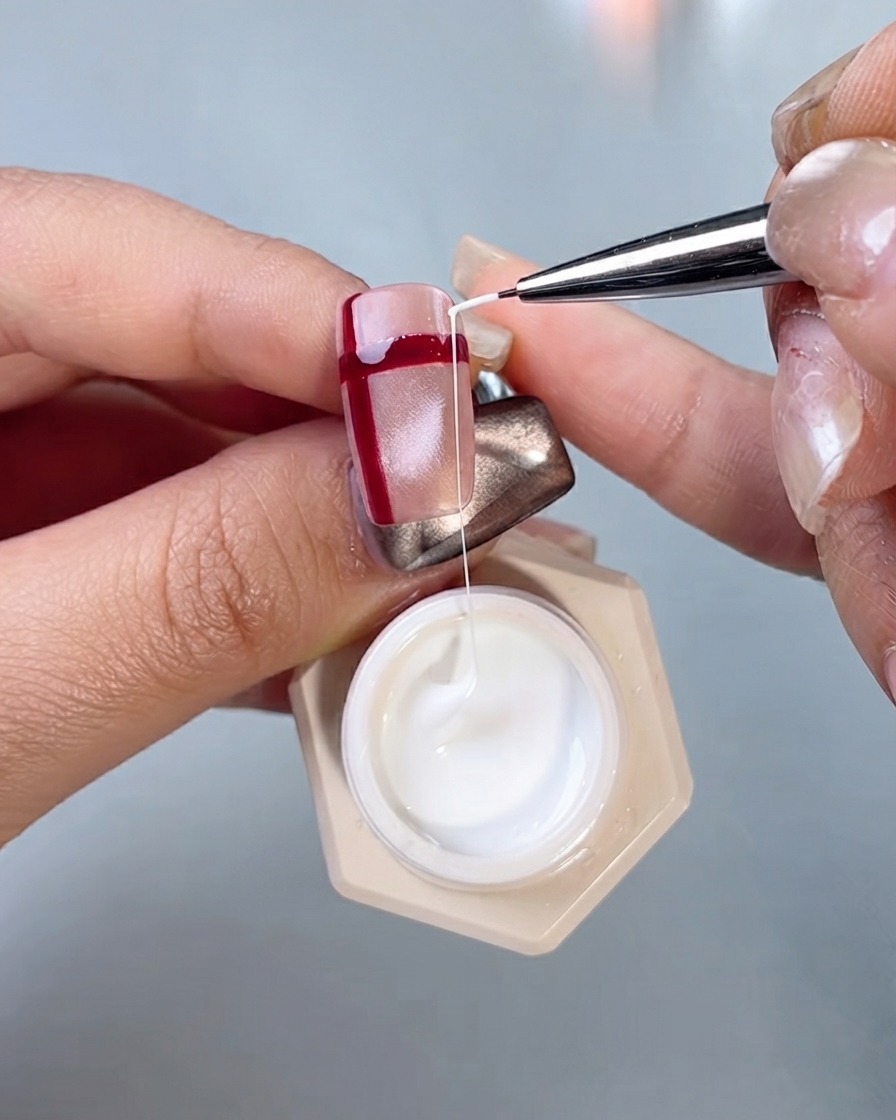 Un pincel de detalle para nail art tirando de una hebra de gel de seda blanco para dibujar líneas cruzadas en una uña con líneas rojas.
