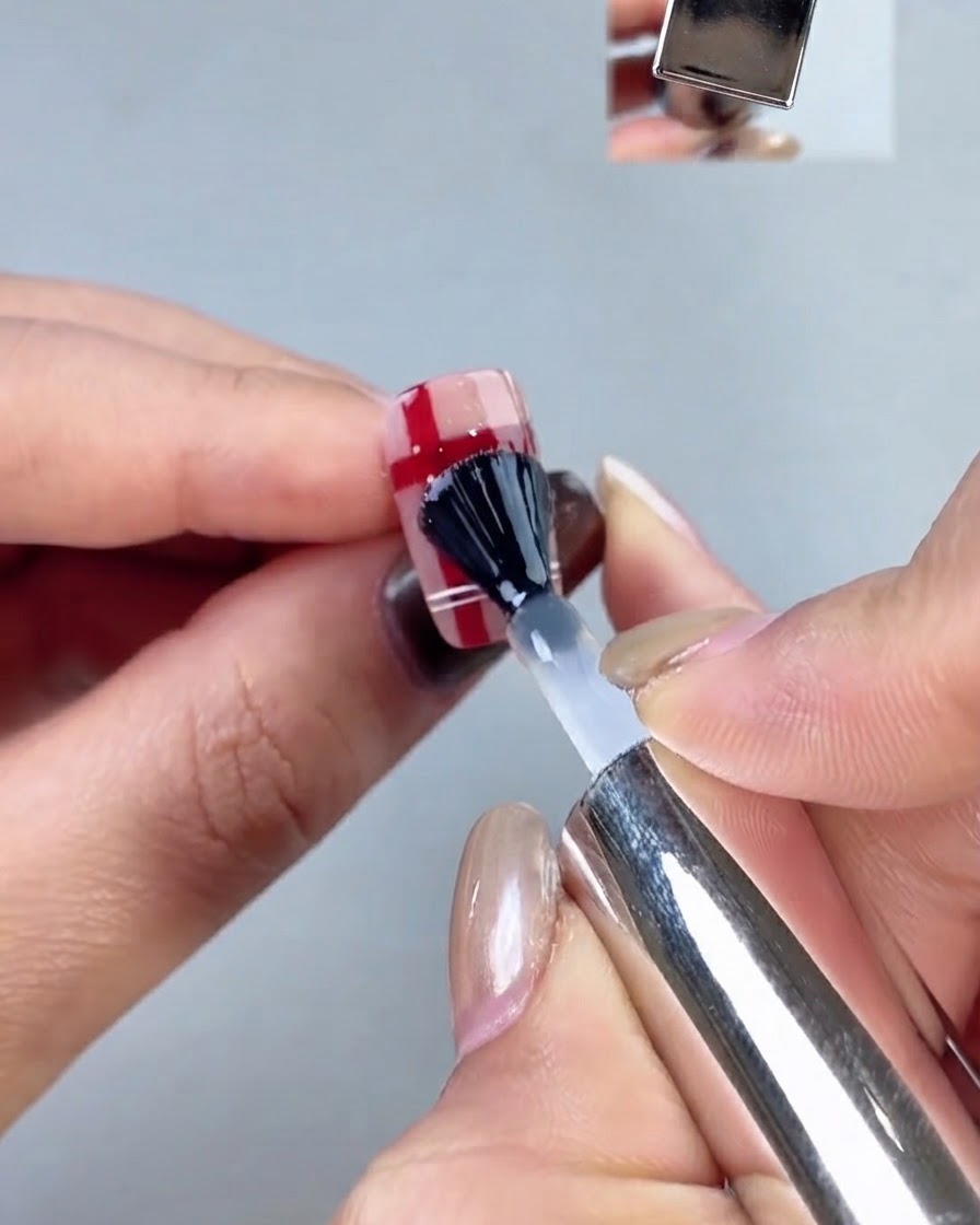 Un pincel de uñas con tapa negra aplicando una capa superior transparente sobre un diseño de uñas a cuadros.