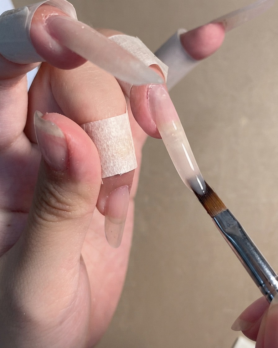 Un pincel de uñas aplicando un gel nude traslúcido sobre una extensión de uña stiletto larga.