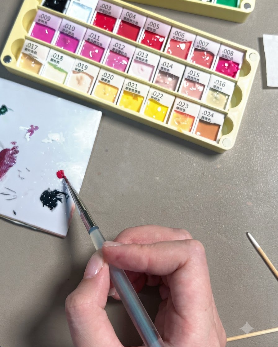 Una mano usando un pincel de detalle fino para depositar un pequeño punto de gel rojo rosa brillante en una paleta de cerámica blanca.