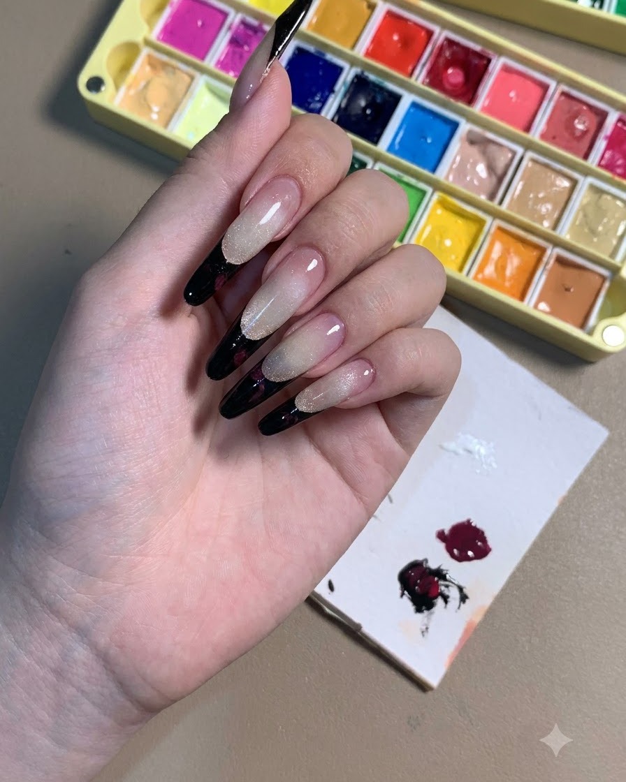Una mano mostrando uñas stiletto con puntas francesas negras y contornos de rosas en tono ciruela oscuro.