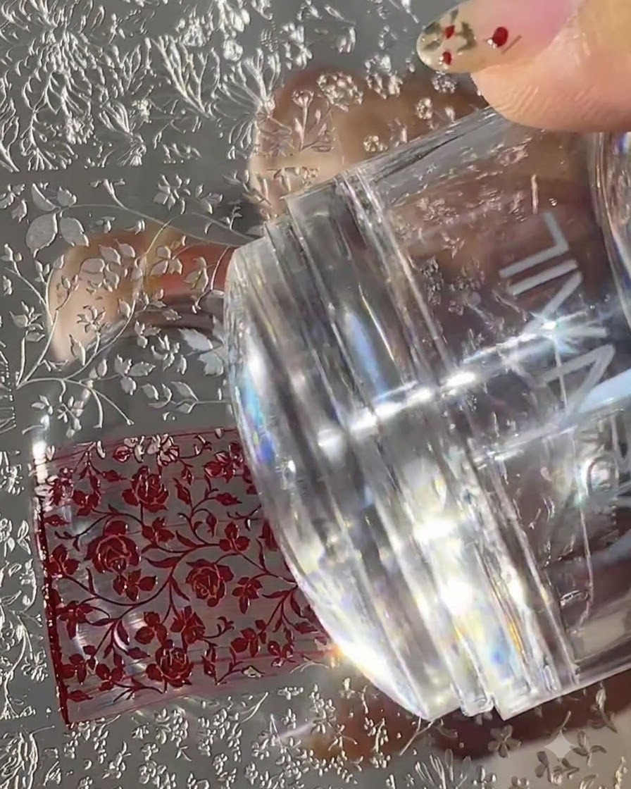 Un sello de uñas de silicona transparente presionando hacia abajo sobre una placa de metal de transferencia grabada llena de patrones florales rojos.