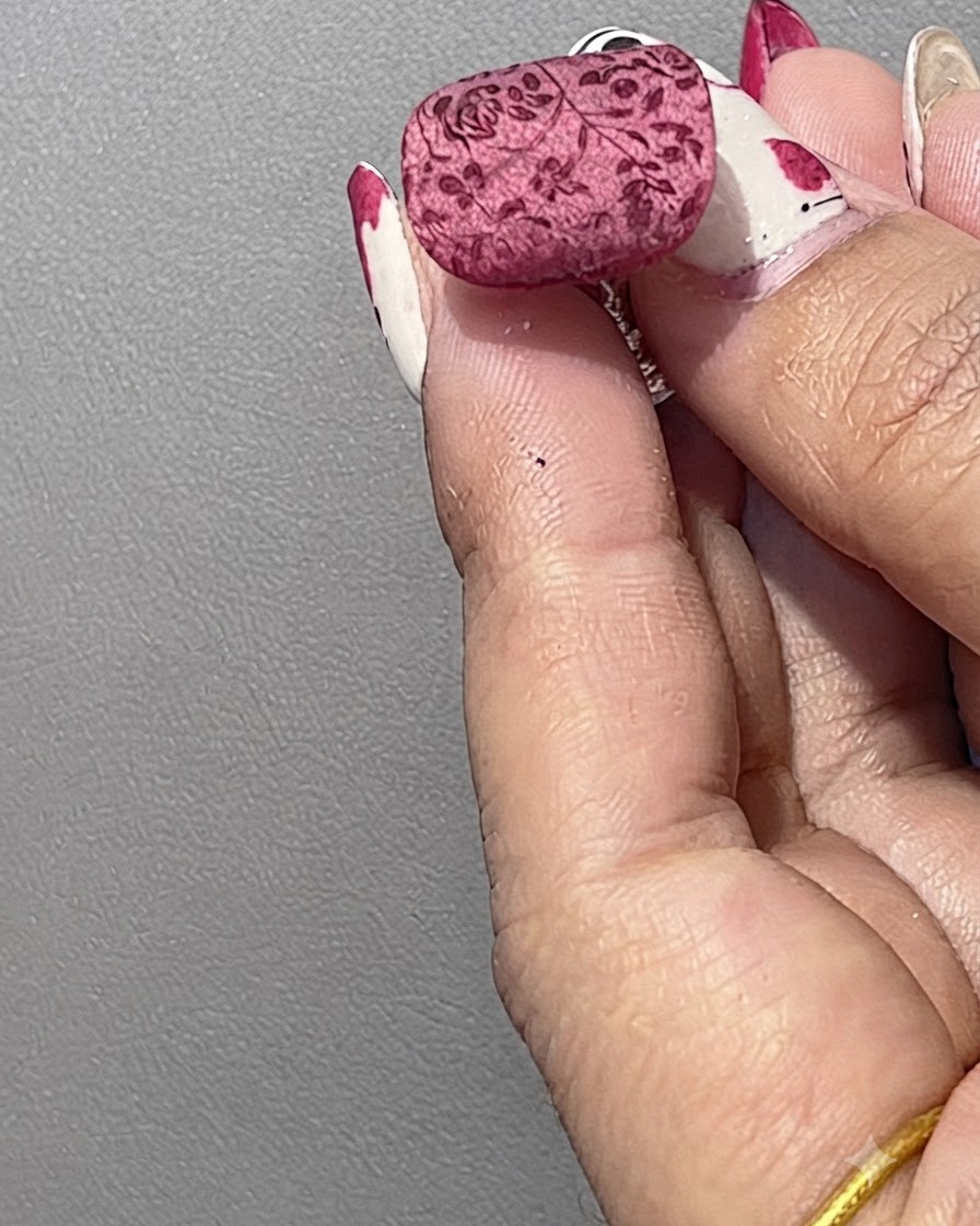 Una uña arreglada con un patrón floral rojo estampado siendo cubierta ligeramente con polvo de cristal blanco usando una espátula.