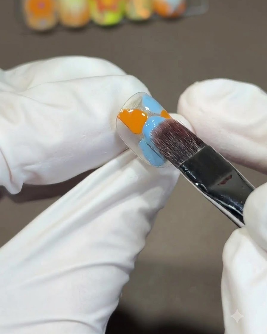 Un pincel cuadrado grande mezclando esmaltes de gel naranja y azul en la punta de una uña usando un trazo descendente en forma de S.