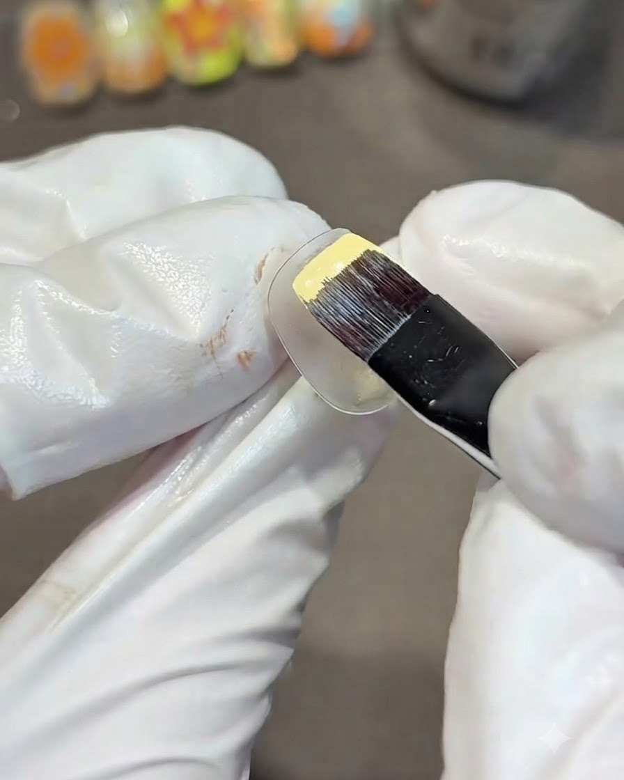 Una mano con guantes usando un pincel de gel plano para pintar una uña de práctica transparente con esmalte de gel amarillo sólido.